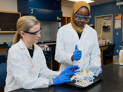 Two scientists working with test tubes in a lab wearing lab coats, safety glasses, and gloves.