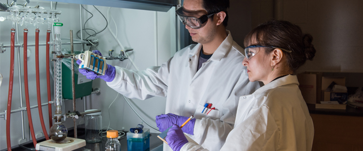 Two scientists working in a lab wearing white lab coats, safety glasses, and gloves.